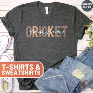 May include: Dark grey heather t-shirt with the text "CRICKET mama" in a layered, distressed font. The text is in shades of brown, pink, and orange. The t-shirt is displayed on a white wooden surface with a pair of white sneakers and a pair of blue jeans.