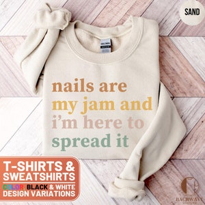 May include: A beige sweatshirt with the text "nails are my jam and i'm here to spread it" in colorful letters. The text is in a retro style font. The sweatshirt is on a wooden surface.