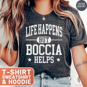May include: Heather dark grey t-shirt with the text "LIFE HAPPENS BUT BOCCIA HELPS" in white. The shirt also has the text "T-SHIRT SWEATSHIRT & HOODIE" in an orange box. The shirt is a casual style.