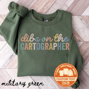 May include: Military green sweatshirt with the text "dibs on the CARTOGRAPHER" in colorful letters. A circular sticker with the words "CUSTOM TEXT" and images of a t-shirt, sweatshirt, and hoodie is on the bottom right.