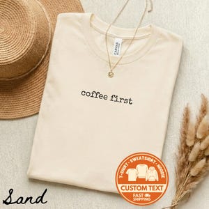 May include: A sand-colored t-shirt with the phrase "coffee first" printed in black. A gold necklace with a small pendant rests on the shirt. A straw hat and dried plants are in the background. A custom text logo is on the bottom right.