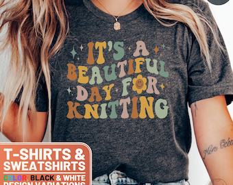 May include: Dark heather grey t-shirt with the text "IT'S A BEAUTIFUL DAY FOR KNITTING" in colorful retro font. The shirt also has a smiling flower graphic. The shirt is part of a collection of t-shirts and sweatshirts.