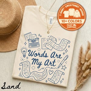 May include: Sand-colored t-shirt with a blue illustration of a typewriter, lightbulb, and flowing text, with the words "Words Are My Art". A round sticker indicates the shirt is available in 10+ colors.
