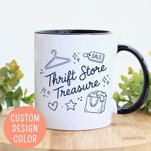 May include: White ceramic mug with a black interior and handle. The mug features the text "Thrift Store Treasure" in blue script, along with illustrations of a hanger, shopping bag, and sale tag. A peach-colored circle with the text "CUSTOM DESIGN COLOR" is on the left.