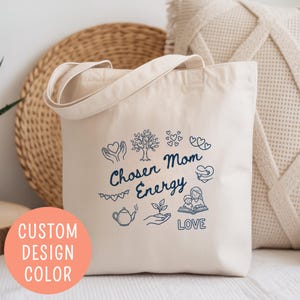 May include: Cream-colored tote bag with the words "Chosen Mom Energy" and various blue illustrations. The bag has a simple design with handles and is made of a soft fabric. A round sticker on the bottom left says "Custom Design Color".