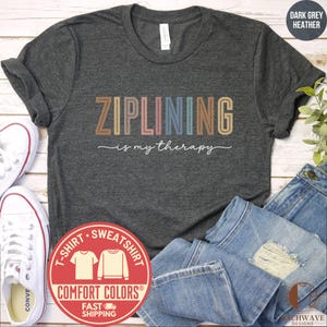 May include: Dark grey heather t-shirt with the text "ZIPLINING is my therapy" in colorful block letters. The shirt is paired with blue jeans and white sneakers. A Comfort Colors label is visible.