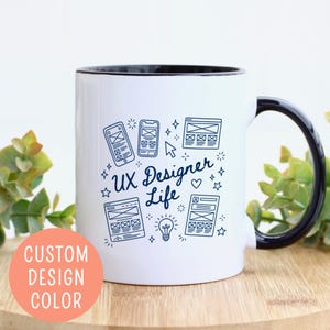 May include: White ceramic mug with a black interior and handle. The mug features a design with the text "UX Designer Life" and illustrations of mobile phones, website layouts, and a lightbulb. A peach-colored circle with the text "CUSTOM DESIGN COLOR" is on the left.