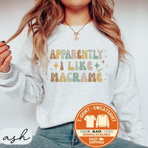 May include: A light gray sweatshirt with the text "APPARENTLY I LIKE MACRAME" in retro-style lettering. The design includes colorful text and star accents. A circular graphic at the bottom right indicates color and design options, and fast shipping.