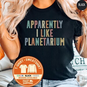 May include: Black t-shirt with the text "APPARENTLY I LIKE PLANETARIUM" in colorful block letters. The shirt has a crew neck and short sleeves. A circular graphic at the bottom indicates the availability of black and white designs, and fast shipping.