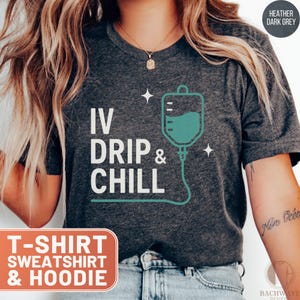 May include: Heather dark grey t-shirt with the text "IV Drip & Chill" and a teal IV drip bag graphic. The shirt also has the text "T-SHIRT SWEATSHIRT & HOODIE" in a rectangular box. The shirt is made of a soft material.