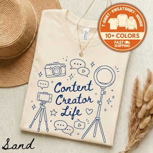 May include: Sand-colored t-shirt with a blue graphic design featuring a camera, speech bubbles, a ring light, and the text "Content Creator Life." The shirt also has a tag that says "Canvas."