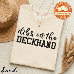 May include: A sand-colored t-shirt with the text "dibs on the DECKHAND" in black. The shirt is displayed with a straw hat, a gold necklace, and dried decorative plants. A circular orange graphic reads "T-SHIRT - SWEATSHIRT - HOODIE CUSTOM TEXT FAST SHIPPING".