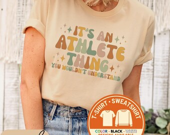 May include: A sand-colored t-shirt with the text "IT'S AN ATHLETE THING YOU WOULDN'T UNDERSTAND" in retro-style lettering. The shirt also features a graphic indicating t-shirt and sweatshirt options, with color choices available.