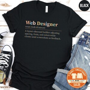 May include: Black t-shirt with the text "Web Designer" in colorful letters. The definition of a web designer is printed below. A pair of jeans and a black and white sneaker are visible in the background.
