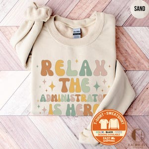 May include: Sand-colored sweatshirt with the text "RELAX THE ADMINISTRATOR IS HERE" in retro-style lettering. The design includes stars and a color palette of brown, yellow, green, and pink. The sweatshirt has a crew neck and long sleeves.