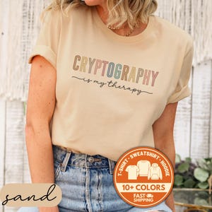 May include: A sand-colored t-shirt with the text "CRYPTOGRAPHY is my therapy" in a colorful, rainbow font. The shirt has rolled-up sleeves and is paired with blue jeans. A circular orange graphic indicates multiple color options and fast shipping.