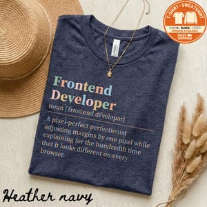 May include: Heather navy t-shirt with the text "Frontend Developer" in colorful letters. The shirt includes a definition and a description of the job. A gold necklace and a straw hat are also in the image.
