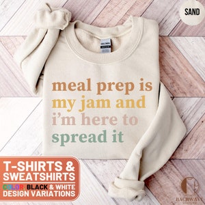 May include: A beige sweatshirt with the text "meal prep is my jam and i'm here to spread it" in rainbow colors. The text is in a retro font style. The sweatshirt is on a wooden background.