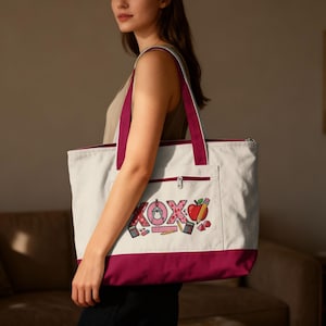 May include: A white canvas tote bag with a burgundy bottom, trim, and straps. The bag features a front pocket and a zipper closure. The front of the bag has a colorful embroidered design with the letters "XOXO", an apple, and school supplies.