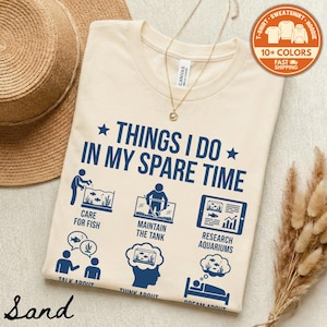 May include: Sand-colored t-shirt with the text "THINGS I DO IN MY SPARE TIME" and illustrations of aquarium-related activities in navy blue. The shirt includes icons for caring for fish, maintaining the tank, and researching aquariums.