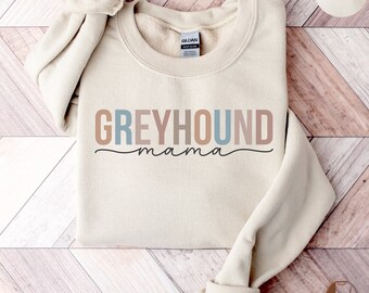 May include: A beige sweatshirt with the text "Greyhound mama" in colorful letters. The sweatshirt is folded and laid on a wooden surface.