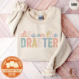 May include: Sand-colored sweatshirt with the text "dibs on the DRAFTER" in pastel colors. The sweatshirt has a crew neck and long sleeves. A circular graphic at the bottom left reads "T-SHIRT - SWEATSHIRT - HOODIE" and "CUSTOM TEXT" with a fast shipping icon.