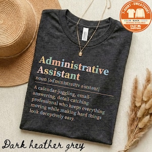 May include: Dark heather grey t-shirt with the words "Administrative Assistant" in colorful letters. The definition of the job is printed below. A gold necklace and a straw hat are also in the image.