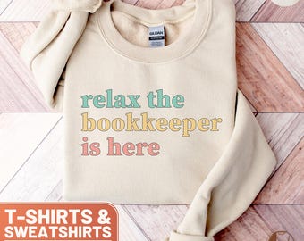 May include: A beige sweatshirt with the text "relax the bookkeeper is here" in pastel colors. The text is in a playful font. The sweatshirt is a crew neck style.