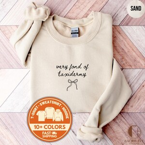May include: Sand-colored sweatshirt with the text "very fond of taxidermy" in black script, along with a bow graphic. The sweatshirt has a crew neck and long sleeves. A circular graphic indicates the availability of the shirt in 10+ colors.