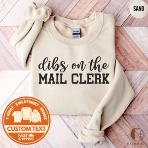 May include: Sand-colored sweatshirt with the text "dibs on the MAIL CLERK" in black. The sweatshirt has a crew neck and long sleeves. A circular graphic with "CUSTOM TEXT" and "FAST SHIPPING" is visible.