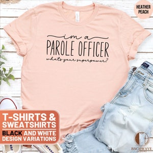 May include: A peach colored t-shirt with the text "I'm a Parole Officer, what's your superpower?" printed in black. The t-shirt is displayed on a wooden surface with a pair of blue jeans and a pair of white sneakers.