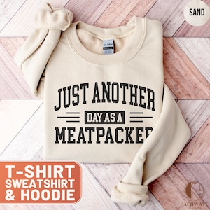 May include: Sand-colored sweatshirt with the text "JUST ANOTHER DAY AS A MEATPACKER" in black. The sweatshirt is folded, with the sleeves visible. The words "T-SHIRT, SWEATSHIRT & HOODIE" are in a red box.