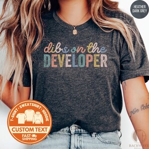 May include: Heather dark grey t-shirt with the text "dibs on the DEVELOPER" in colorful letters. The shirt has a round neck and short sleeves. A circular sticker with the words "CUSTOM TEXT" and "FAST SHIPPING" is visible.