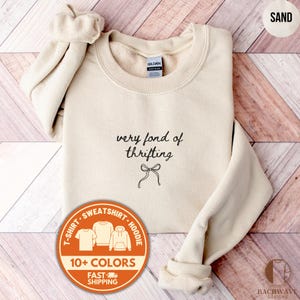 May include: Sand-colored sweatshirt with the text "very fond of thrifting" and a bow graphic. The image includes a circular graphic with shirt styles and the text "10+ COLORS" and "FAST SHIPPING".