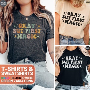 May include: Three t-shirts with the text "Okay but first magic" in a retro font with colorful accents. The shirts are shown in dark grey heather, cream, and black.