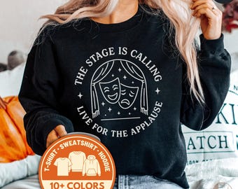 May include: Black sweatshirt with a white graphic featuring theater masks and the text "THE STAGE IS CALLING LIVE FOR THE APPLAUSE." A round sticker indicates the availability of 10+ colors and fast shipping. The sweatshirt is a t-shirt, sweatshirt, or hoodie.