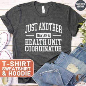 May include: Dark grey heather t-shirt with the text "JUST ANOTHER DAY AS A HEALTH UNIT COORDINATOR" in white. The image also includes a pair of white sneakers, blue jeans, and a sign that says "T-SHIRT, SWEATSHIRT & HOODIE."