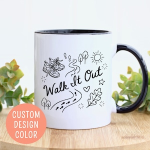 May include: White ceramic mug with a black interior and handle. The mug features a black and white illustration with the text "Walk It Out" and images of sneakers, a path, trees, and a sun. A peach-colored circle with the text "CUSTOM DESIGN COLOR" is on the left.