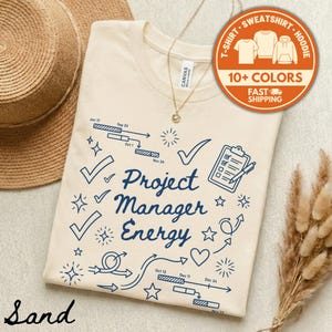 May include: A sand-colored t-shirt with the words "Project Manager Energy" in navy blue, along with checkmarks, stars, and arrows. The shirt also features a graphic with the text "10+ COLORS" and "FAST SHIPPING".
