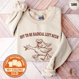 May include: Sand-colored sweatshirt with the text "OFF TO BE RADICAL LEFT SCUM" and a cartoon illustration of a frog riding a goose. The sweatshirt also has a round orange sticker with the text "T-SHIRT SWEATSHIRT HOODIE 10+ COLORS FAST SHIPPING".