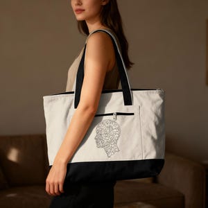 May include: A large tote bag with a white body, black bottom, and black straps. The bag features a black and white line drawing of a human head with a brain-like design on the front pocket. The bag has a zipper closure.
