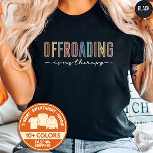 May include: Black t-shirt with the text "OFFROADING is my therapy" in a colorful, retro font. The shirt also features a round orange sticker with the text "10+ COLORS" and images of a t-shirt, sweatshirt, and hoodie.