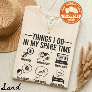 May include: A sand-colored t-shirt with the phrase "THINGS I DO IN MY SPARE TIME" and illustrations of bug-related activities. The shirt includes icons for finding, watching, and studying bugs, as well as talking, thinking, and dreaming about them.