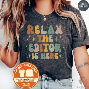 May include: Dark heather grey t-shirt with the text "RELAX THE EDITOR IS HERE" in retro-style lettering. The design includes stars and a color palette of brown, pink, green, and yellow. The shirt also has a graphic indicating t-shirt and sweatshirt options.