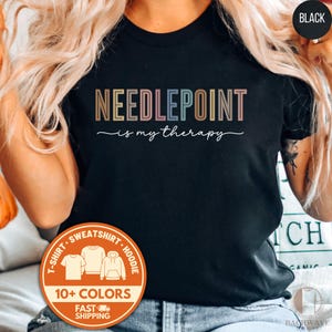 May include: Black t-shirt with the text "NEEDLEPOINT is my therapy" in colorful letters. A circular orange sticker indicates the shirt is available in 10+ colors. The shirt is a t-shirt, sweatshirt, or hoodie.