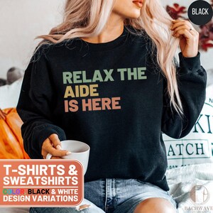May include: Black sweatshirt with the text "Relax the Aide is Here" in a retro font with green, yellow, and orange colors.