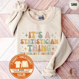 May include: Sand-colored sweatshirt with the text "IT'S A STATISTICIAN THING YOU WOULDN'T UNDERSTAND" in retro-style lettering. The sweatshirt has a crew neck and long sleeves. A circular graphic indicates color and design options.