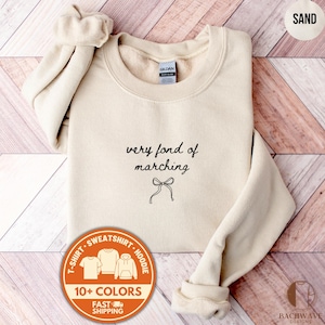 Puede incluir: Sudadera color arena con el texto "very fond of marching" y un gráfico de lazo. La sudadera tiene cuello redondo y mangas largas. Un gráfico circular indica la disponibilidad del diseño en camisetas, sudaderas y sudaderas con capucha en más de 10 colores.