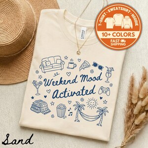 May include: Sand-colored t-shirt with a blue graphic design featuring a sofa, coffee cup, pizza slice, sunglasses, and the text "Weekend Mood Activated." The shirt also has a graphic of a hammock, palm trees, and a sun.