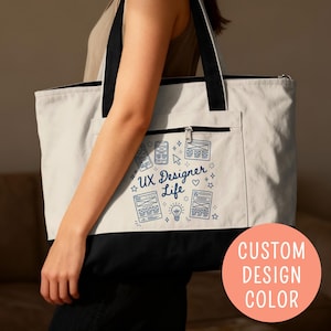 May include: A white tote bag with a black bottom and straps. The front pocket features a blue graphic design with the text "UX Designer Life" and related icons. The bag has a zipper closure and a peach-colored circle with the text "CUSTOM DESIGN COLOR".